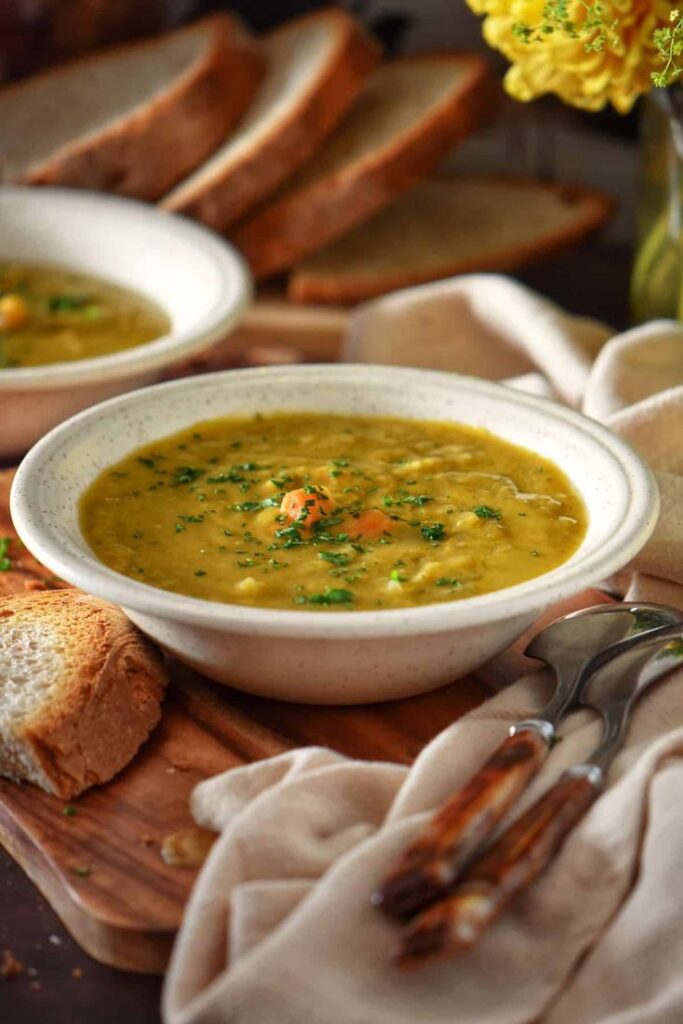 Bowl of hearty vegetable soup garnished with herbs, served with fresh crusty bread on a wooden board.