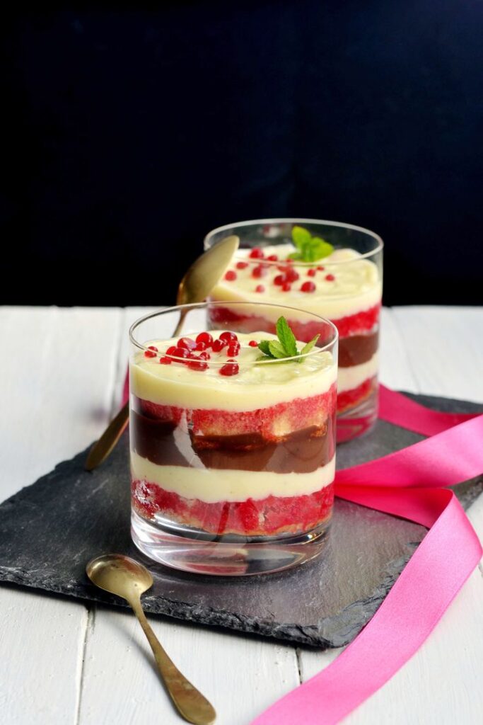 Layered dessert in glasses with red sponge, cream, chocolate, and pomegranate seeds, garnished with mint leaves.
