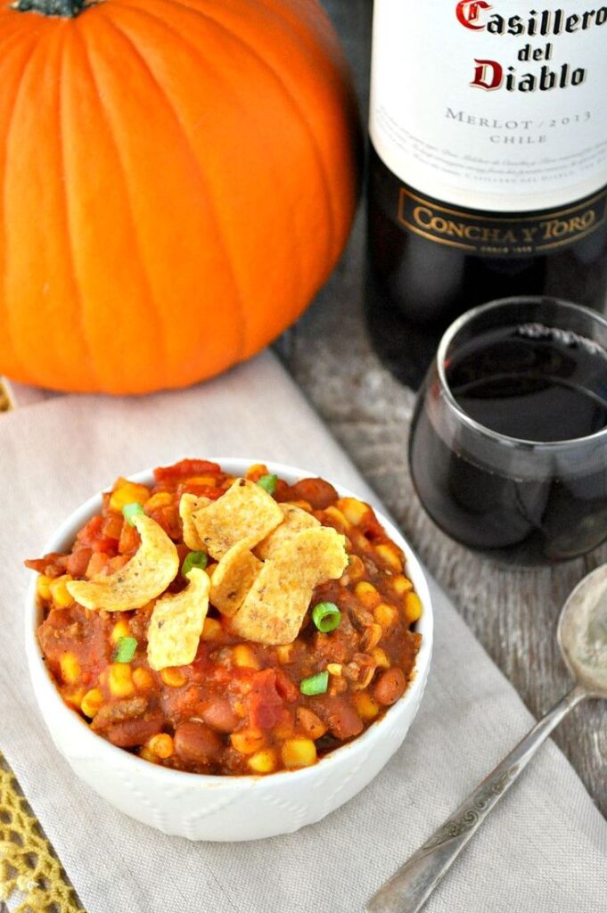 Bowl of chili with corn chips and green onions beside pumpkin and wine, perfect autumn meal setting.