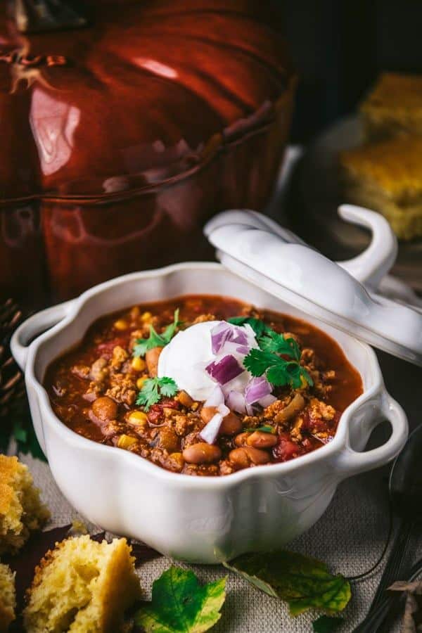 Hearty chili in a white bowl garnished with sour cream, red onions, and cilantro beside cornbread. Perfect comfort food.