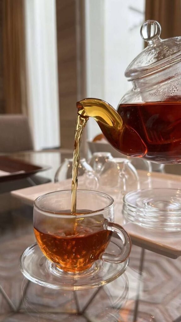 Glass teapot pouring hot tea into a clear glass cup on a saucer, set on a reflective table.