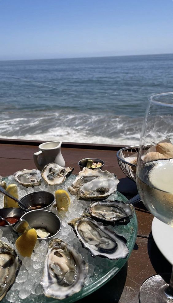 Plate of fresh oysters with condiments by the seaside, served with white wine and bread, under a clear blue sky.