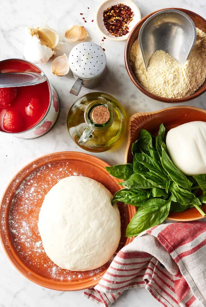 margherita-pizza-recipes - Master the Art of Homemade Pizza with Fun, Simple Steps | Pizza ingredients on a table: dough, basil, olive oil, canned tomatoes, garlic, and spices.