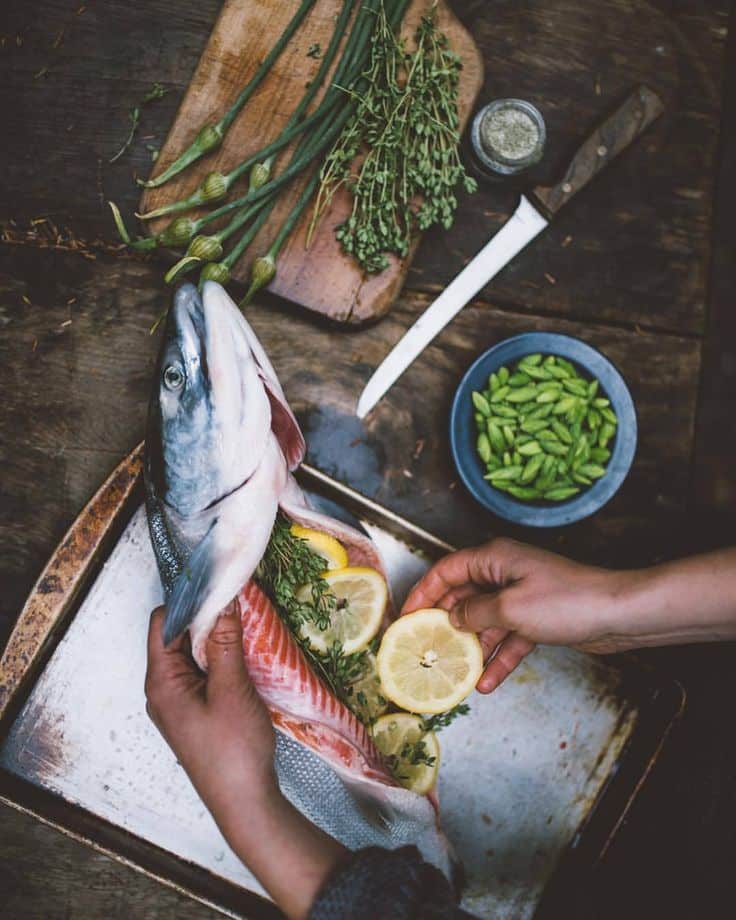Person preparing stuffed fish with herbs and lemon, surrounded by fresh ingredients on a rustic table.