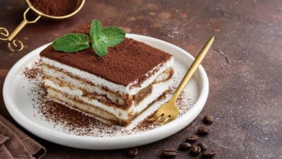 Delicious tiramisu dessert with cocoa topping and mint garnish on a white plate beside a golden fork.