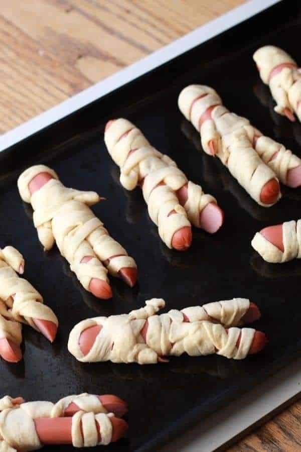 Halloween mummy hot dogs wrapped in pastry on a baking sheet, ready for serving as a spooky snack.