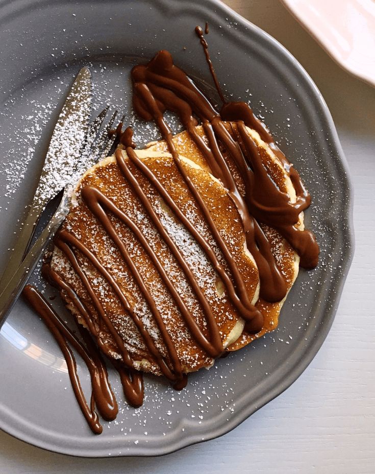 Delicious pancakes topped with chocolate sauce and powdered sugar on a gray plate, with fork and knife on the side.