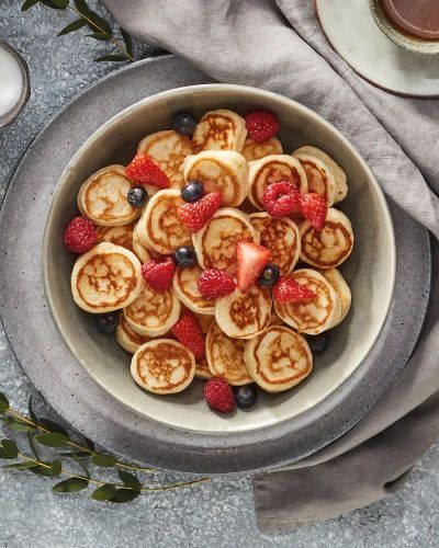 A bowl of mini pancakes topped with fresh raspberries, strawberries, and blueberries, served on a gray tablecloth.