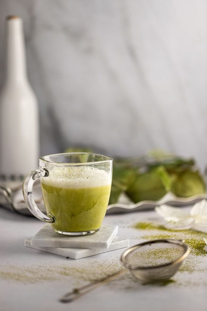 Clear glass mug of frothy green matcha latte on marble coasters with sifter nearby.