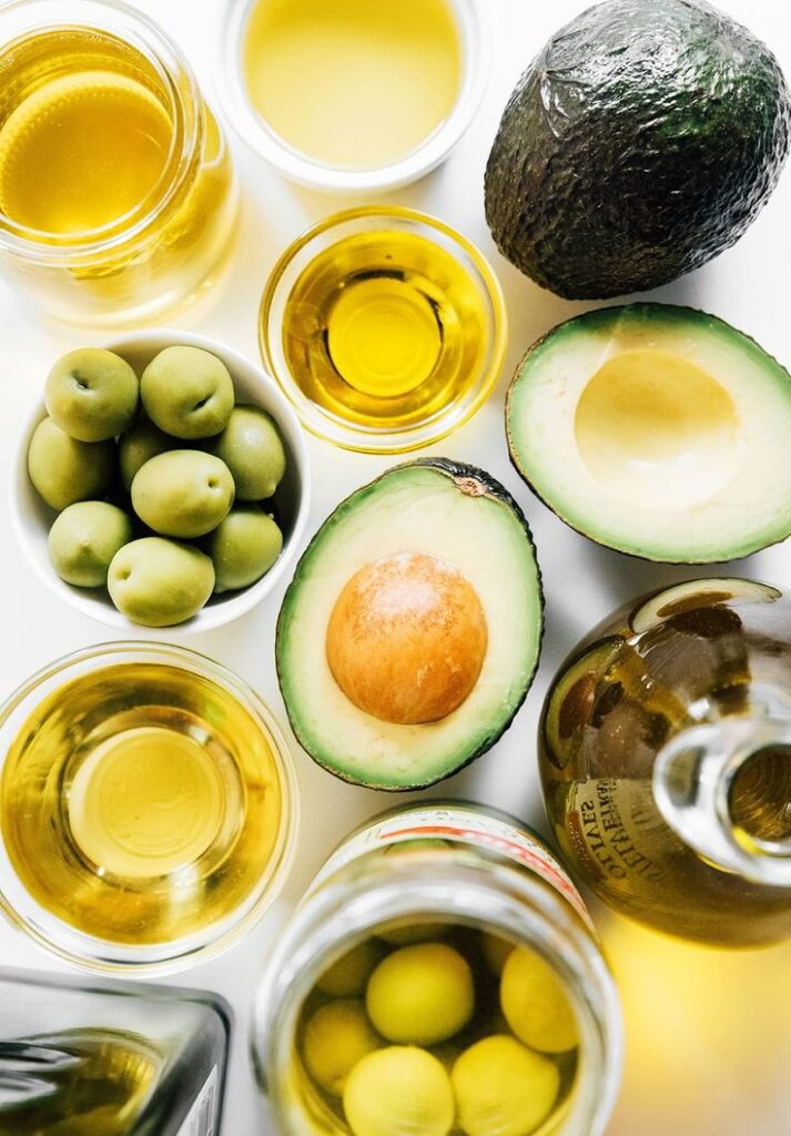 Avocado, olives, and various oils in jars on a white background, emphasizing healthy fats and Mediterranean diet.