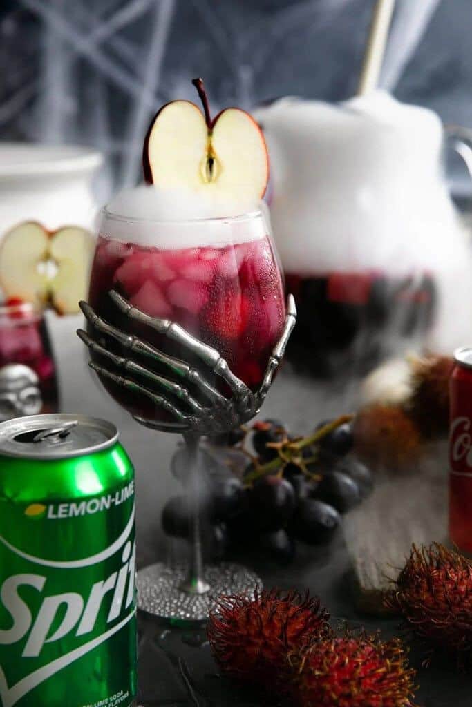 Halloween-themed cocktail with apple garnish in skeleton glass, surrounded by mist, Sprite, grapes, and rambutans.