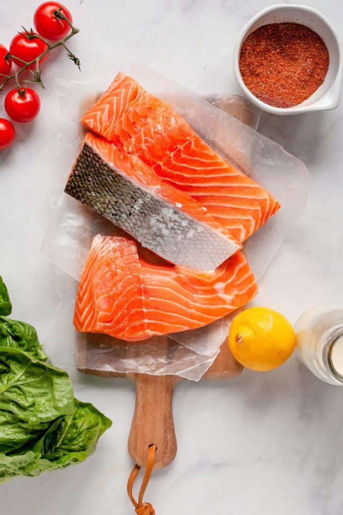Fresh salmon fillets on a cutting board with tomatoes, lemon, lettuce, and spices on a white background.