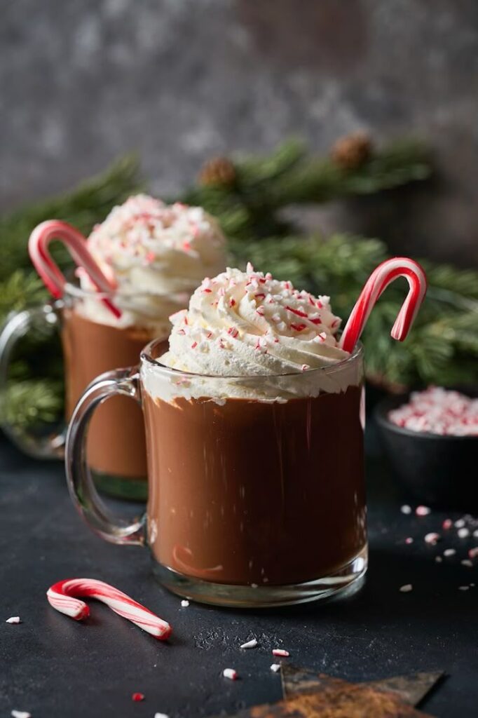 Two mugs of hot chocolate topped with whipped cream and candy canes, surrounded by festive greenery and crushed candy.