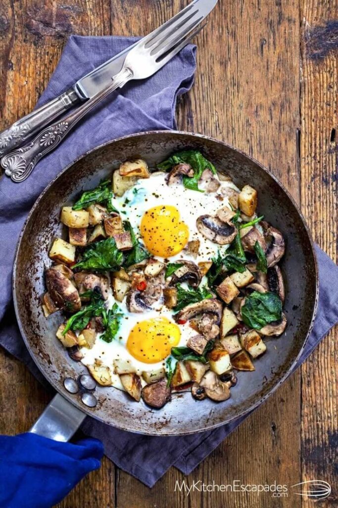 Skillet with fried eggs, potatoes, mushrooms, and greens on a wooden table, garnished with black pepper.