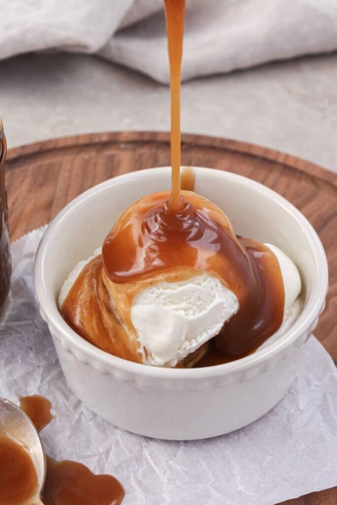 Caramel sauce drizzling over vanilla ice cream in a white bowl, on a wooden tray background.