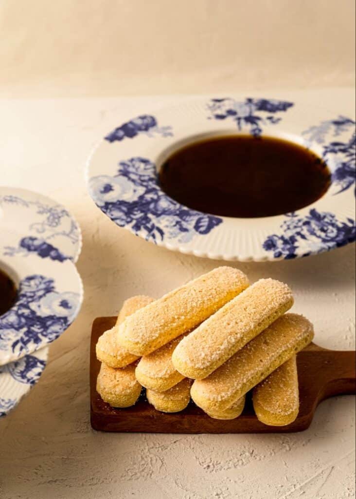 Ladyfingers on a wooden board with floral cups of coffee, set against a light background. Perfect for a dessert setting.