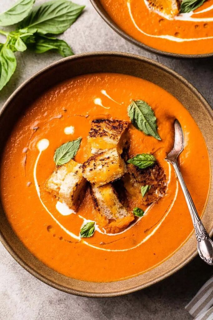 Creamy tomato soup topped with grilled cheese croutons and fresh basil in a bowl with a spoon.