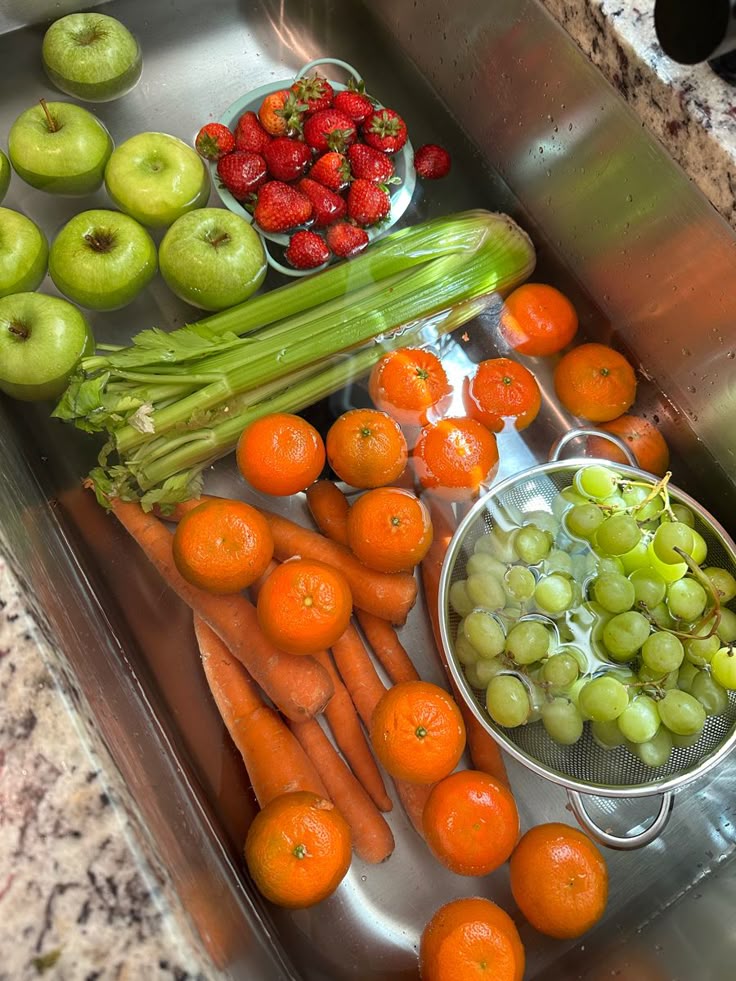 Fresh fruits and vegetables soaking in a kitchen sink, including apples, strawberries, celery, oranges, and grapes.