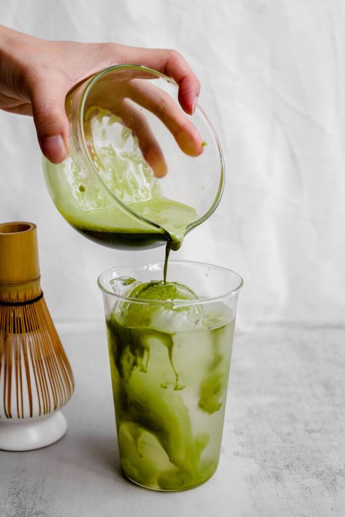 Pouring fresh matcha over ice cream in a clear glass, with a bamboo whisk nearby.