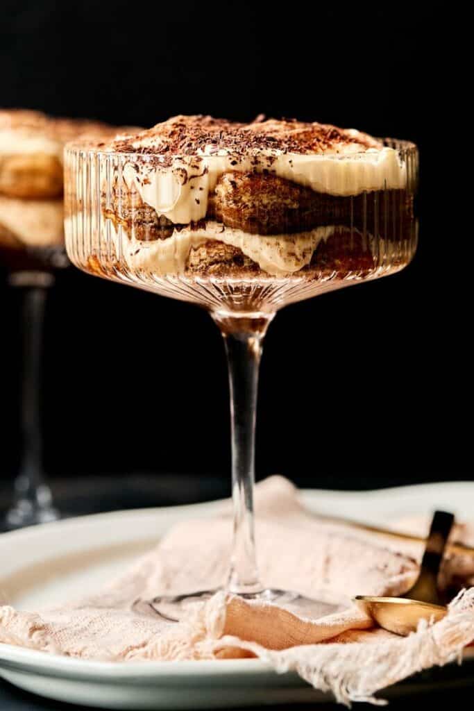 Decadent tiramisu in an elegant glass, layered with creamy mascarpone and dusted with cocoa, on a white plate.
