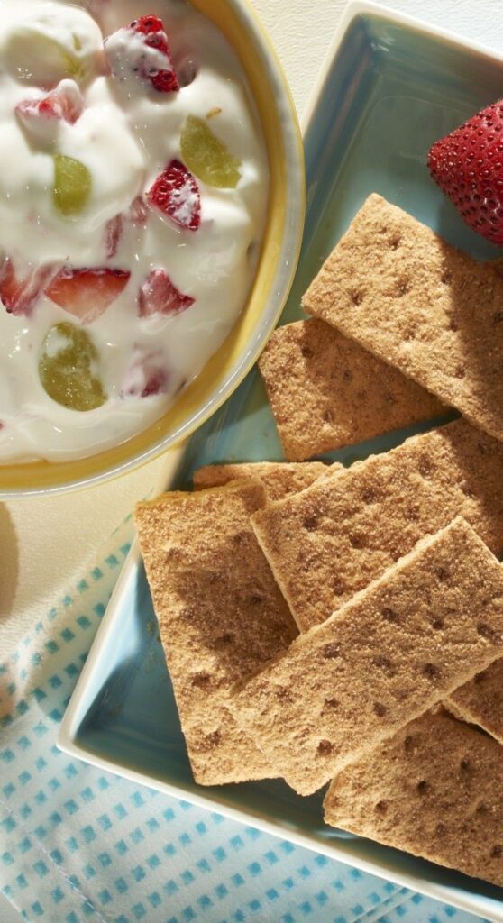 Bowl of yogurt with strawberries and grapes beside crunchy graham crackers on a plate, perfect breakfast snack.
