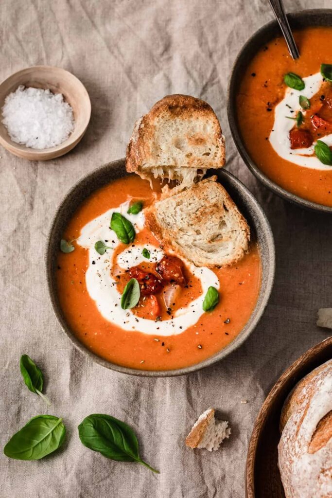 Creamy tomato soup garnished with basil and served with crusty bread on a cloth background.