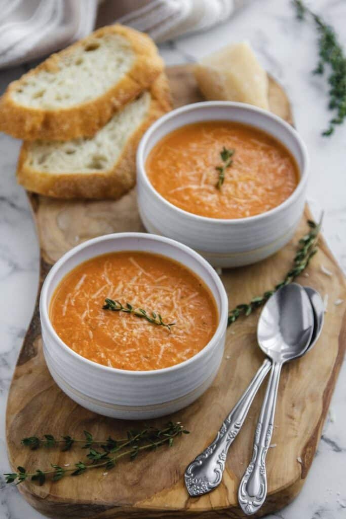 Two bowls of tomato soup garnished with thyme, served with crusty bread on a wooden board.