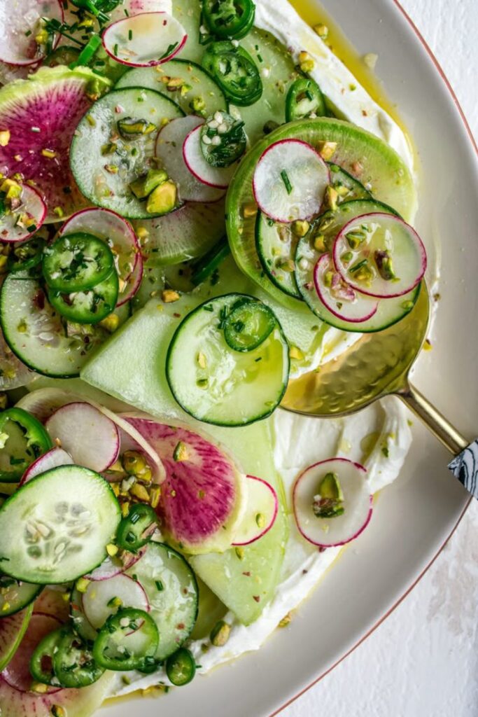Fresh salad with cucumber, radish, jalapeño, and pistachios on a plate, topped with herbs and olive oil.