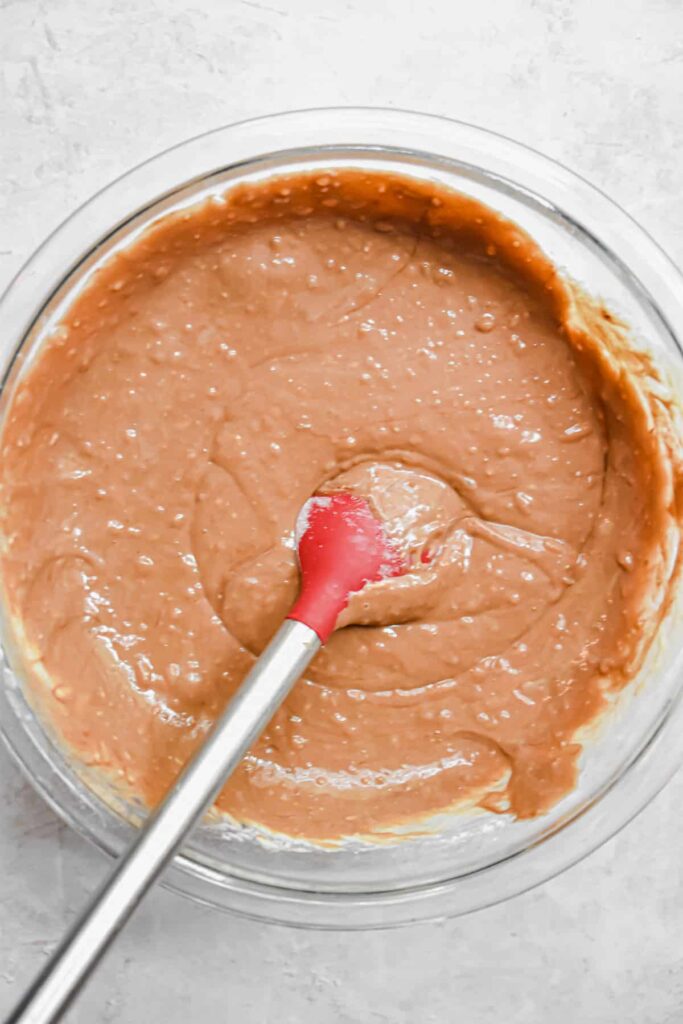 Chocolate cake batter in glass bowl with red spatula, ready for baking. Rich and creamy mixture on a light surface.