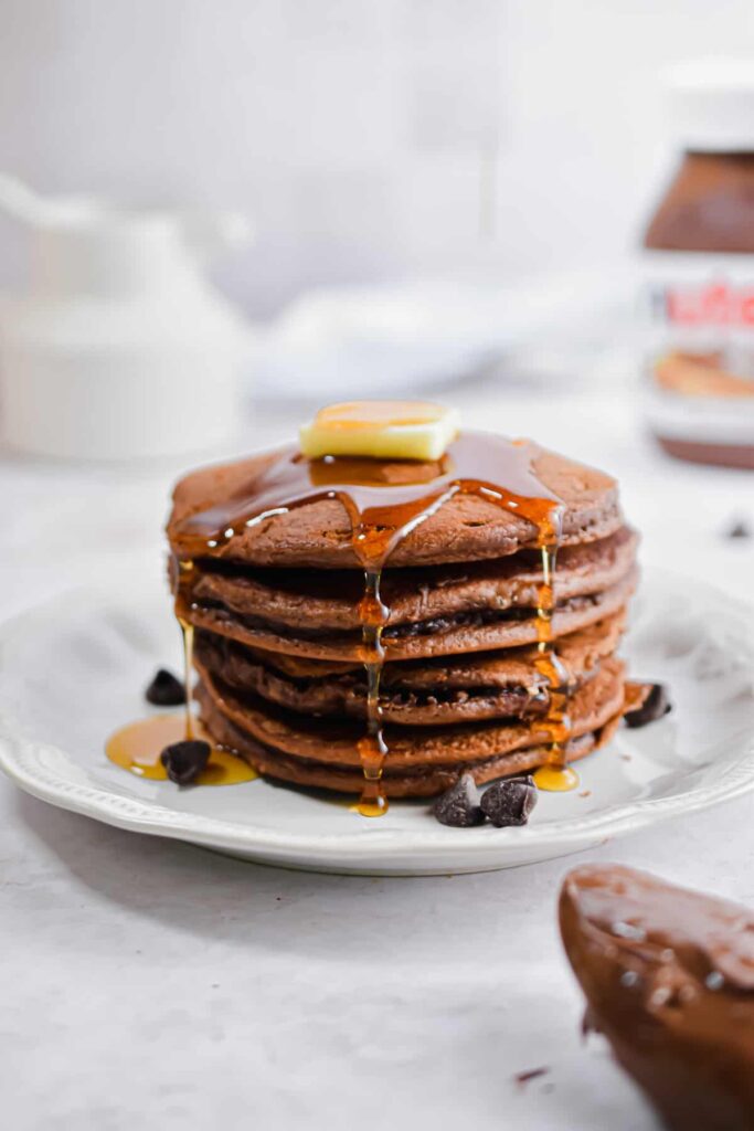 Stack of chocolate pancakes with syrup and butter on top, surrounded by chocolate chips on a white plate.