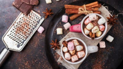 Cups of hot chocolate topped with colorful marshmallows, surrounded by cinnamon and chocolate shavings.
