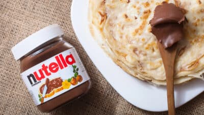 Jar of Nutella and crepes on a plate with a wooden spoon, on a burlap surface.
