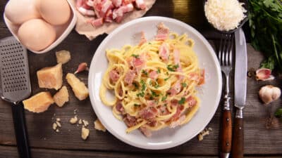 Plate of creamy spaghetti carbonara with parmesan, bacon, eggs, and herbs on a rustic wooden table.