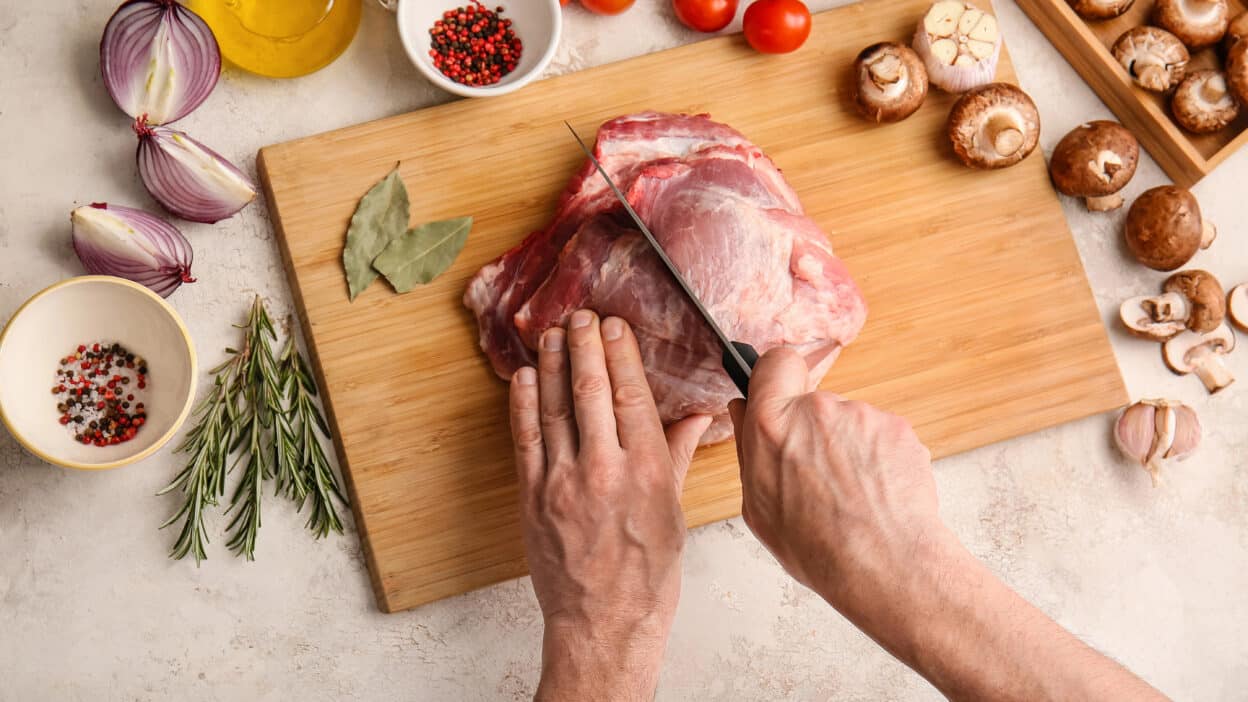 Cutting raw meat on a wooden board with herbs, mushrooms, onions, and spices for cooking preparation.