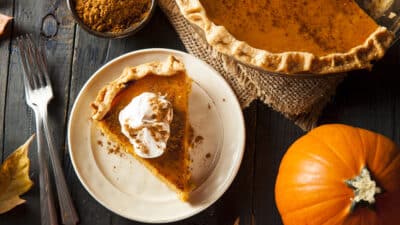 Slice of pumpkin pie with whipped cream on plate, surrounded by autumn leaves and pumpkin on rustic table.