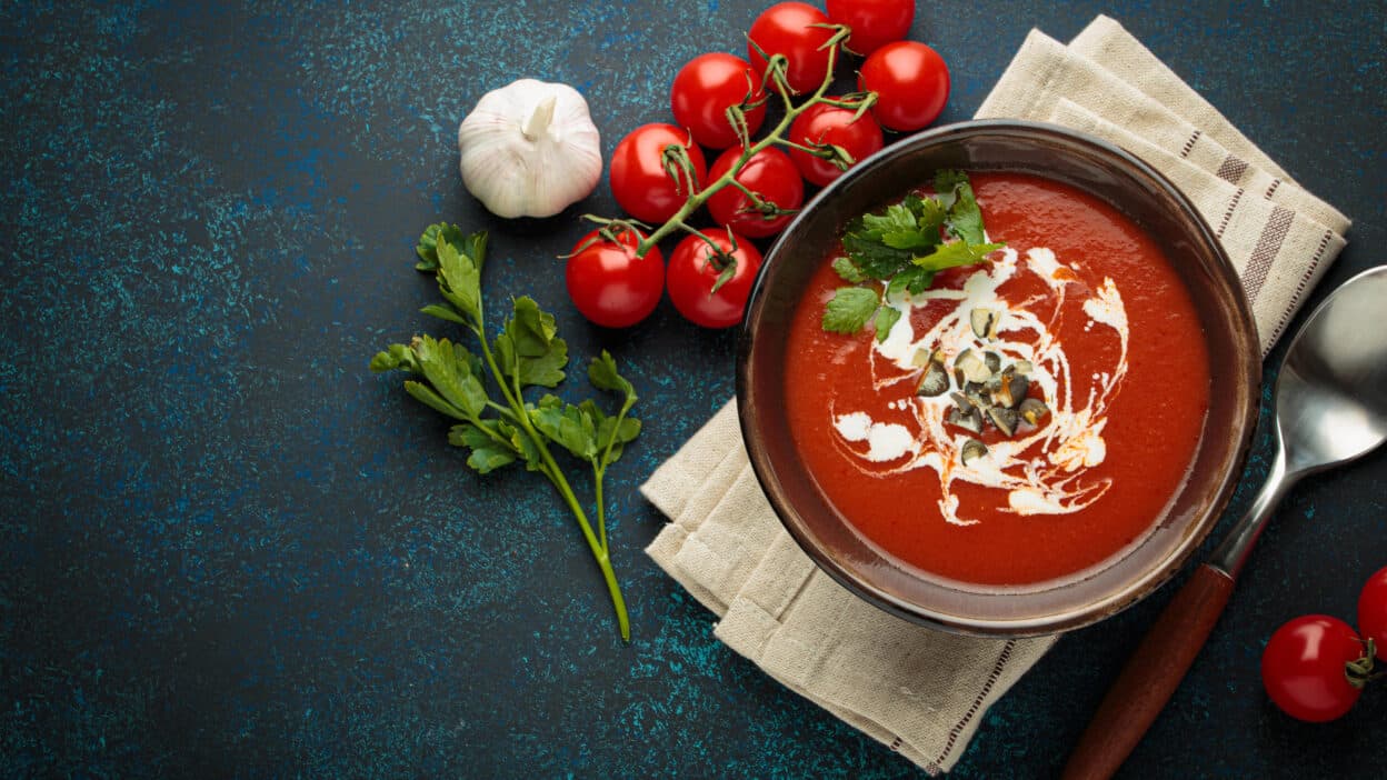 Bowl of creamy tomato soup garnished with herbs and olives, surrounded by fresh tomatoes, garlic, and parsley.