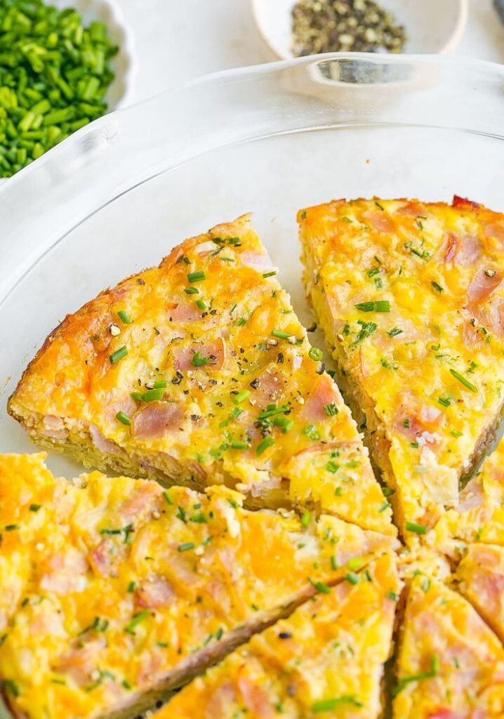 Cheesy ham and chive quiche slices in a clear pie dish, garnished with fresh herbs.