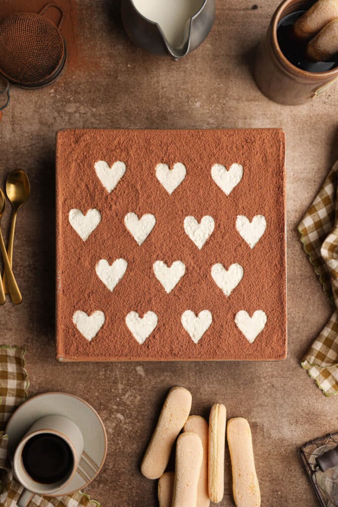 Heart-patterned tiramisu with cocoa topping, surrounded by coffee and ladyfingers on a rustic table setting.