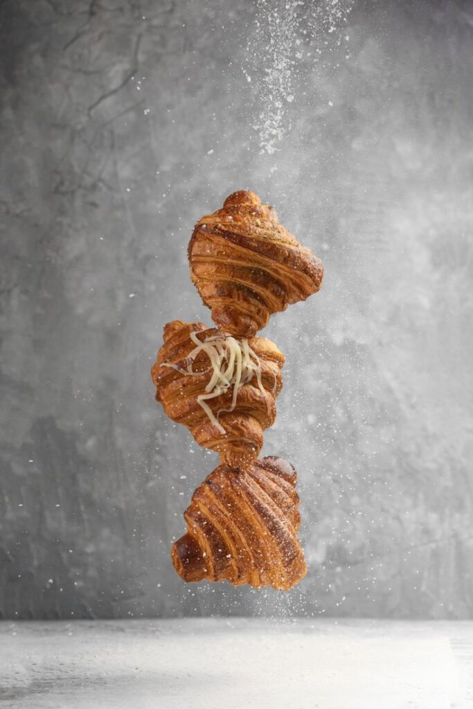Flaky croissants suspended mid-air with a sprinkling of flour on a gray background.