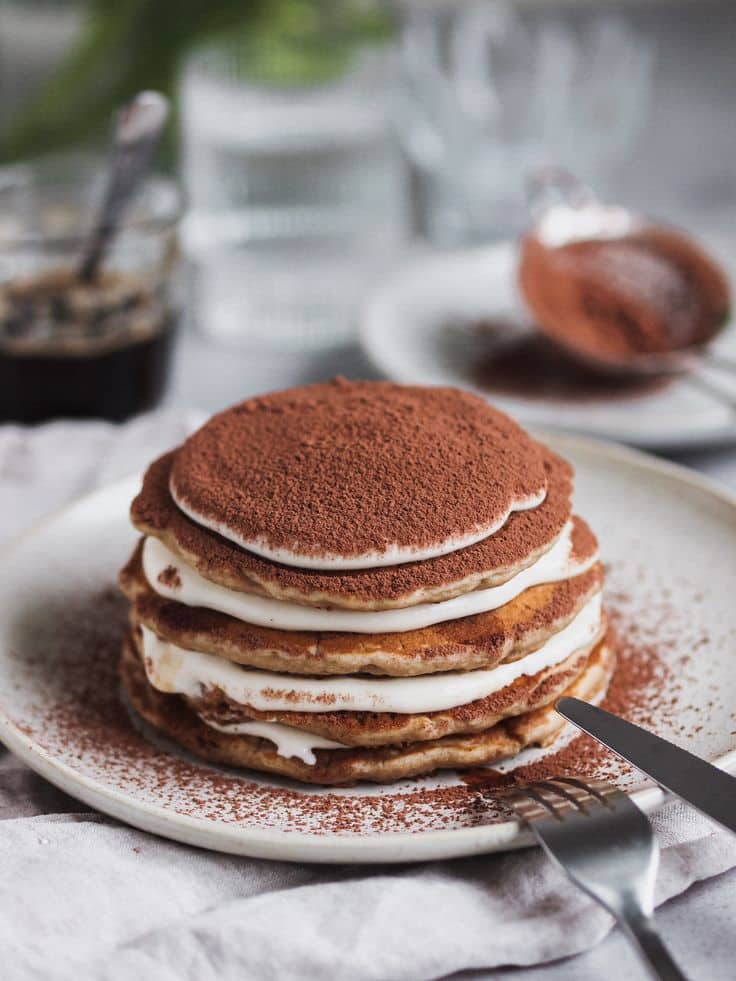Delicious stack of chocolate pancakes with cream layers dusted with cocoa on a white plate.