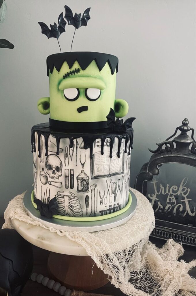Halloween-themed cake with Frankenstein topper, black bats, spooky decorations, and skull details on a white base.