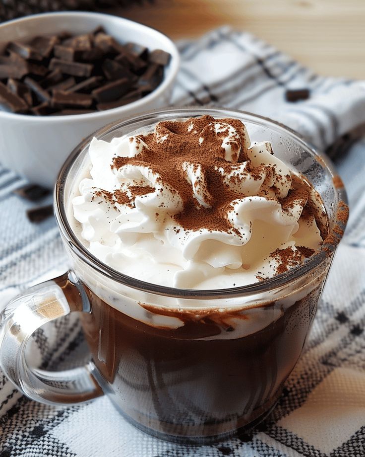 Hot chocolate with whipped cream and cocoa powder in a glass mug, with chocolate pieces in a white bowl behind.