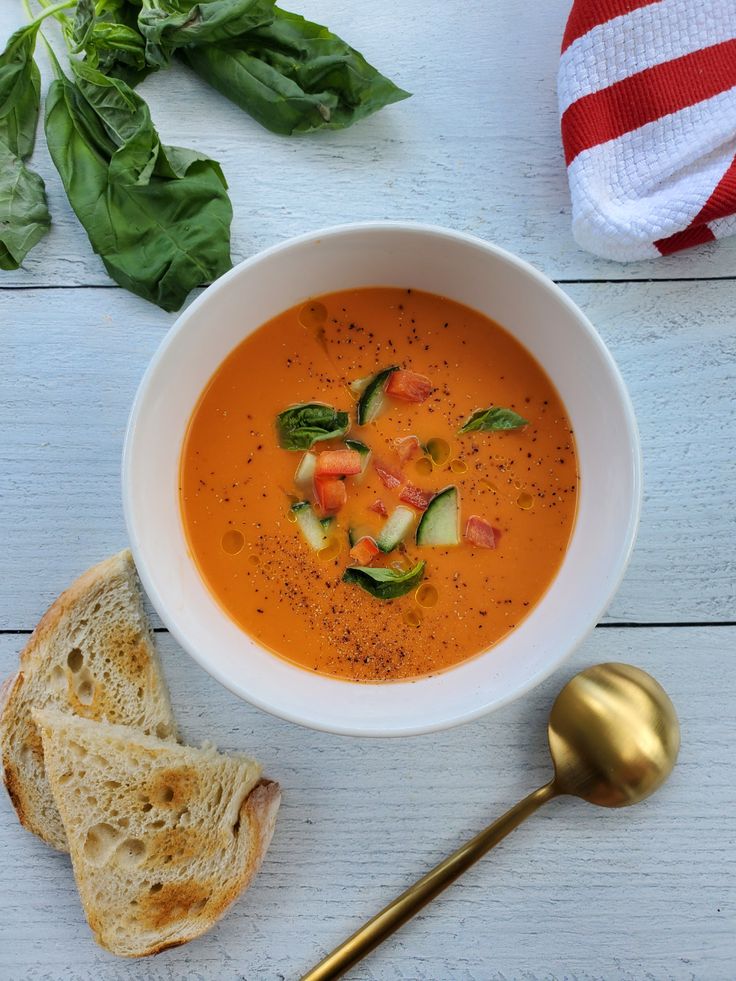 Bowl of tomato soup with basil, cucumber, and pepper garnishes, served with toast and a gold spoon on a white table.
