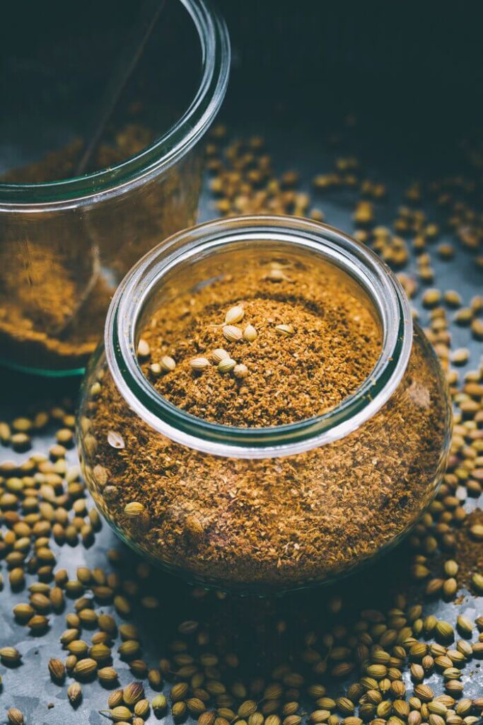Glass jar filled with ground coriander seeds, surrounded by loose seeds on a dark surface.