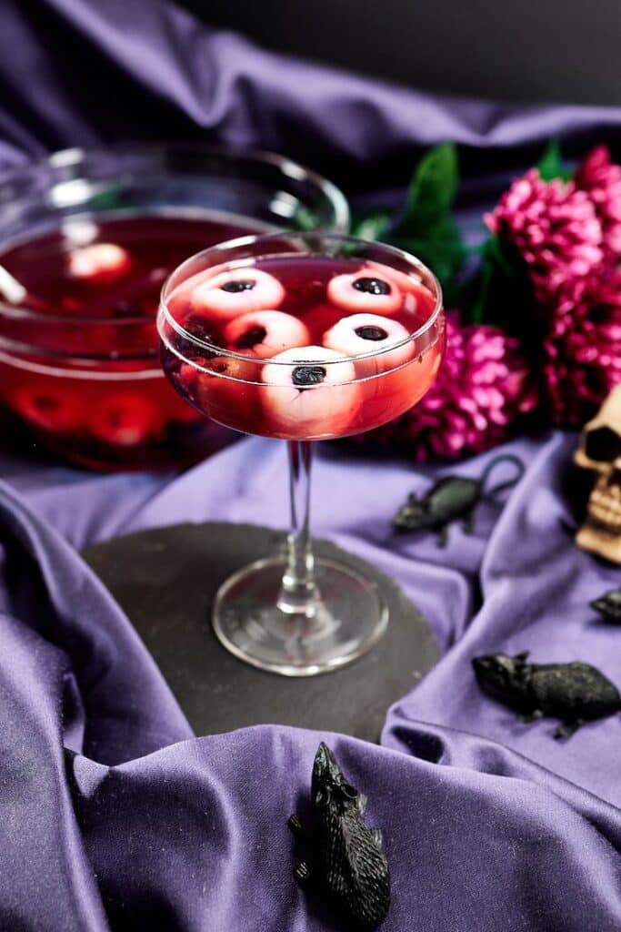 Spooky Halloween cocktail with floating eyeballs, surrounded by creepy decor on a purple fabric background.