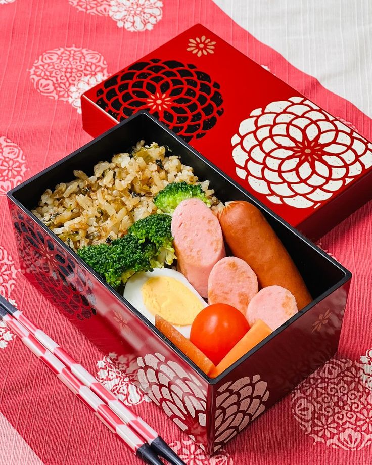 Colorful bento box with rice, sausages, egg, broccoli, and cherry tomatoes, served on a decorative red cloth.