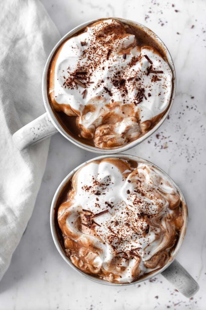 Two mugs of rich hot chocolate topped with whipped cream and chocolate shavings on a marble surface.