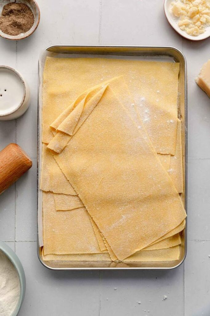 Fresh homemade pasta sheets on a tray, surrounded by ingredients, ready for making lasagna.