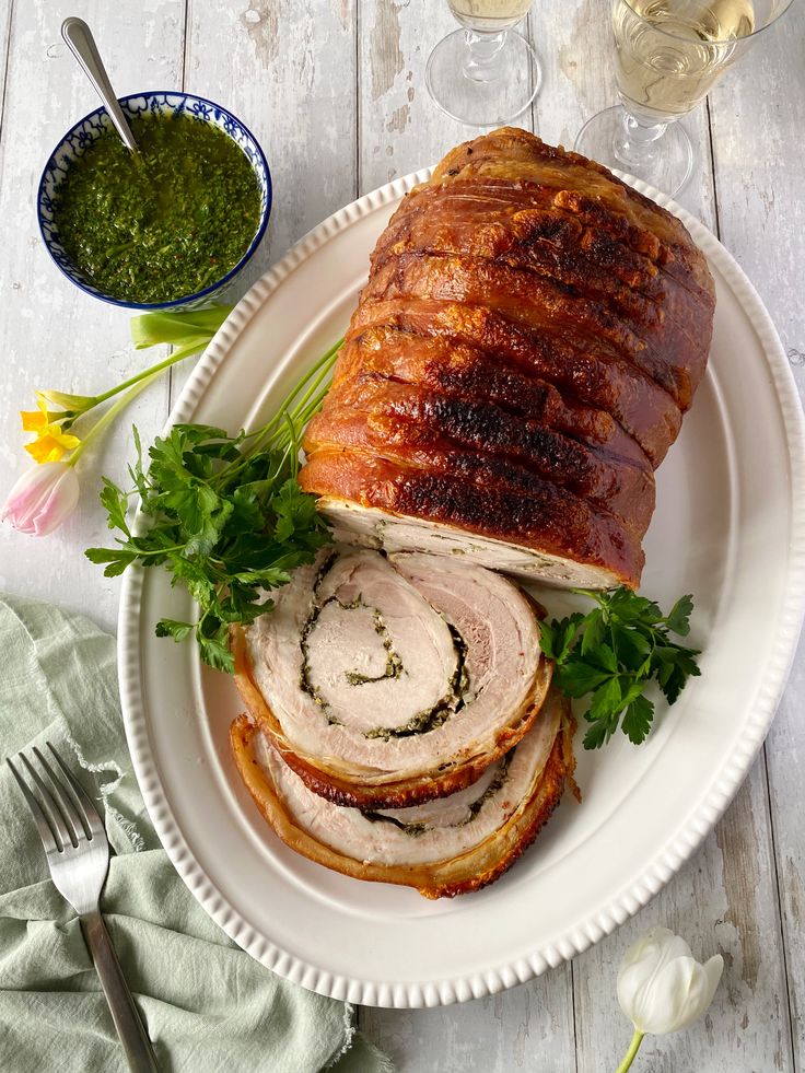 Porchetta roast sliced on a platter with fresh herbs, served with chimichurri sauce, wine glasses in the background.