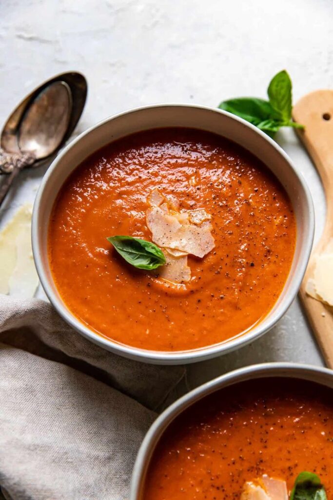 Creamy tomato soup garnished with Parmesan and basil in a white bowl on a table with a spoon nearby.